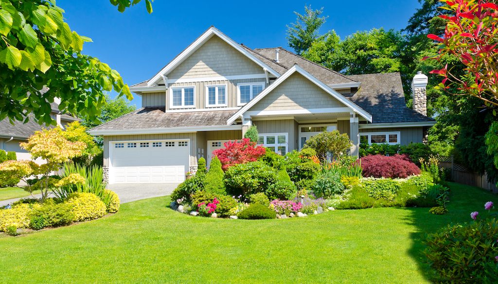 A picturesque two-story house with a well-manicured front lawn and colorful garden showcases expert landscaping in Simpsonville. The home features a gray exterior, white trim, and steep gables. The lush garden includes various shrubs, flowers, and trees, creating a vibrant and welcoming atmosphere.